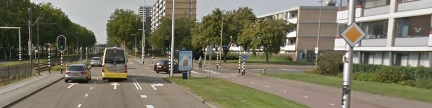 Een straatbeeld van de kruising Beneluxlaan met de Struyckenlaan. Aan de linkerkant is de trambaan en rijden auto's en een bus die de kruising oversteken. Er zijn twee rijstroken voor rechtdoorgaand verkeer en een rijstrook voor rechtsaf richting de Struyckenlaan. Aan de rechterkant staan hoge appartementsgebouwen, en aan de rechterkant is het fietspad langs de weg te zien. Bomen en groen vullen de achtergrond van de straat.