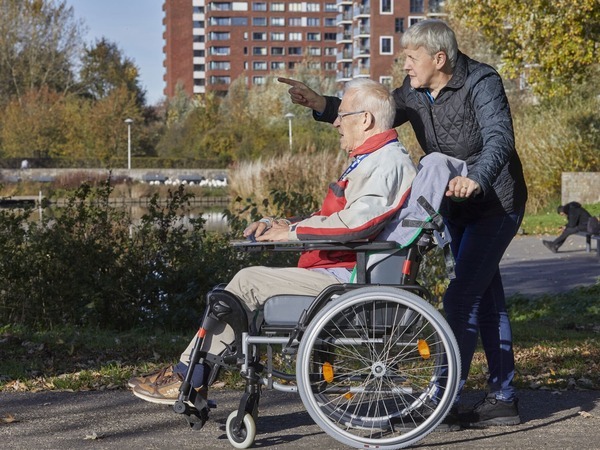 Een vrouw begeleidt een oudere man in een rolstoel over een pad in een groene omgeving.